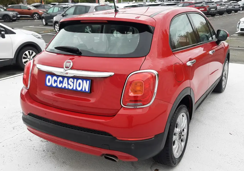 Vue 3/4 arrière droite d'une FIAT 500X rouge avec plaque "OCCASION" sur parking extérieur.