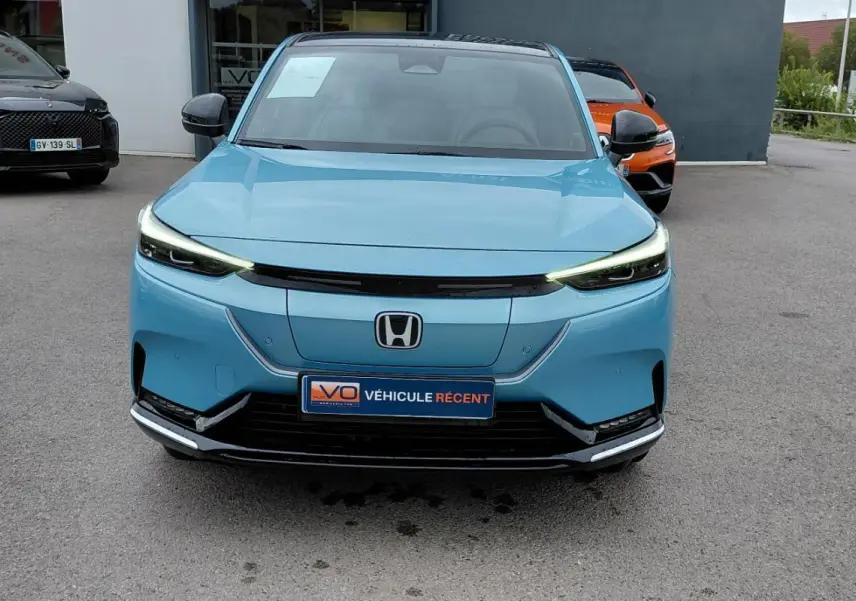 Vue frontale d'une Honda e:Ny1 Advance 204 ch 2025 en bleu électrique avec phares LED allumés sur parking.