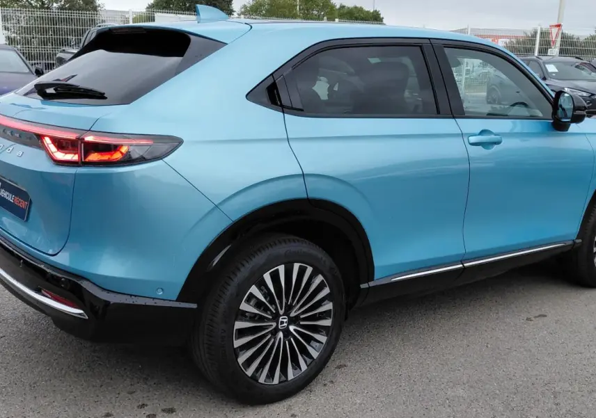 Vue 3/4 arrière droite d'un Honda e:Ny1 Advance bleu électrique 2025 avec feux arrière allumés et jantes stylisées.