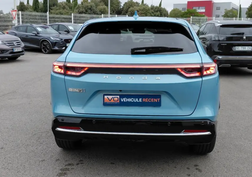 Vue arrière d'un Honda e:Ny1 bleu 2025 avec feux LED allumés et logo Honda visible sur un parking.
