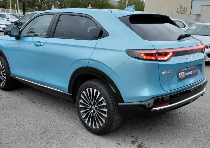 Vue 3/4 arrière droite d’un Honda e:Ny1 bleu électrique 2025 avec feux arrière allumés et jantes stylisées.