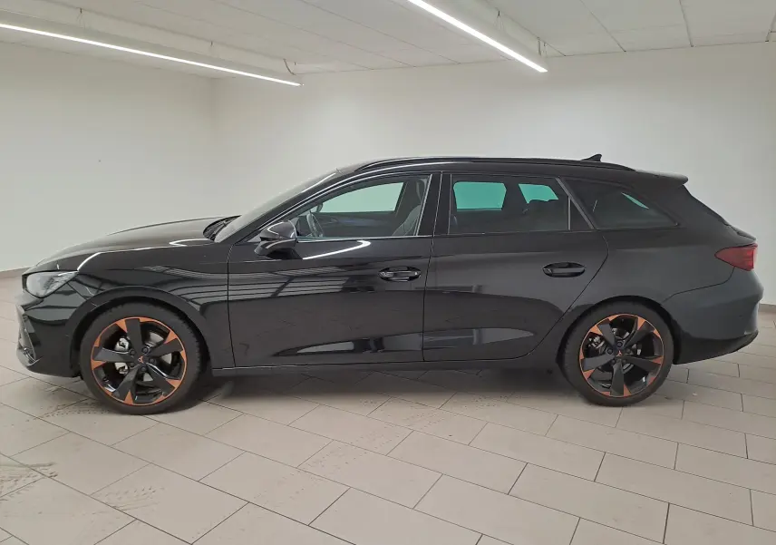 Vue de profil côté gauche d'une CUPRA LEON ST 1.5 eTSI noire avec jantes alliage 18'' cuivrées dans un intérieur lumineux.