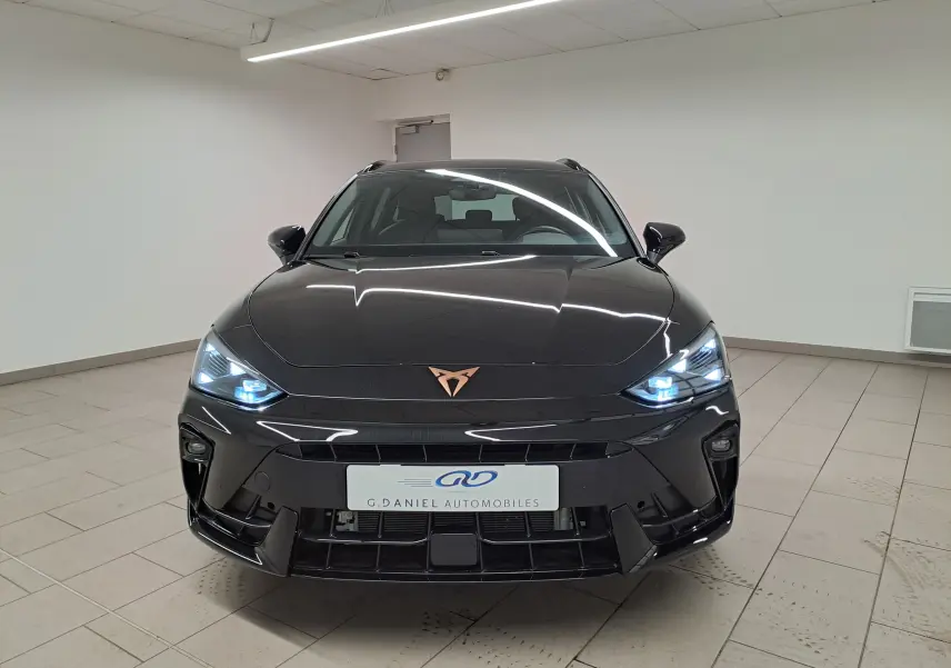Vue frontale d'une CUPRA LEON ST 1.5 eTSI 150 DSG7 en Noir Minuit avec phares allumés et logo cuivré distinctif.