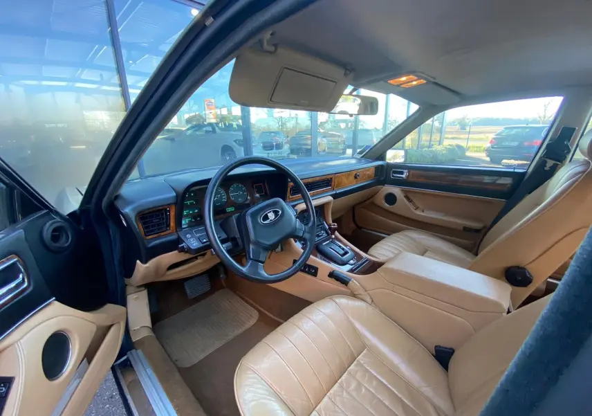 Vue intérieure côté conducteur d'une Jaguar XJ6 3.6 Sovereign 1987 bleu, avec sièges cuir beige et tableau de bord bois.