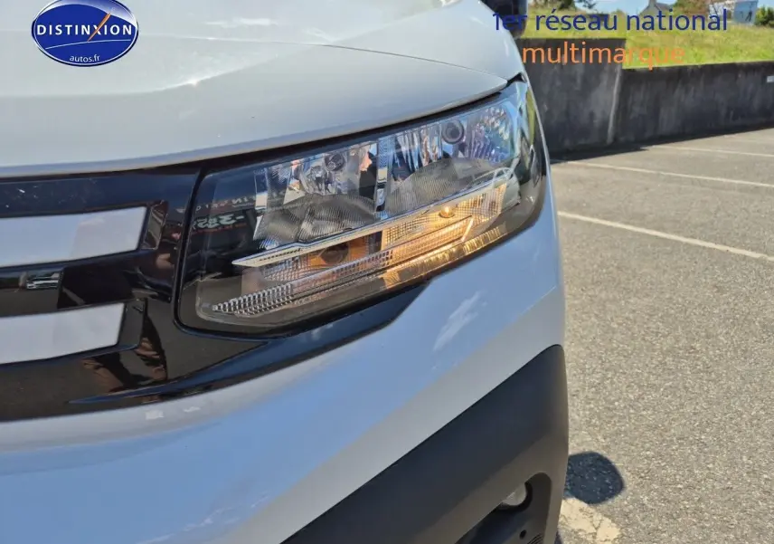 Gros plan sur le phare avant droit du Peugeot Partner 2025 blanc, avec calandre noire et détails lumineux allumés.