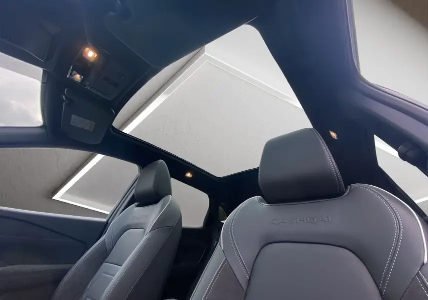 Vue intérieure du Nissan Qashqai 2025 montrant les sièges avant noirs avec surpiqûres blanches et toit panoramique.