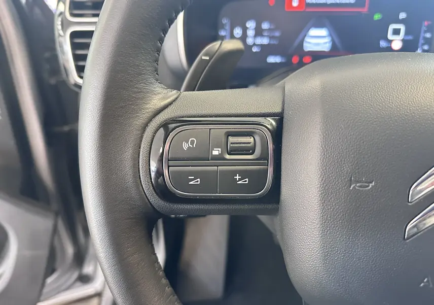 Gros plan sur les commandes gauche du volant cuir noir du Citroën C5 Aircross 2024, avec écran numérique en arrière-plan.