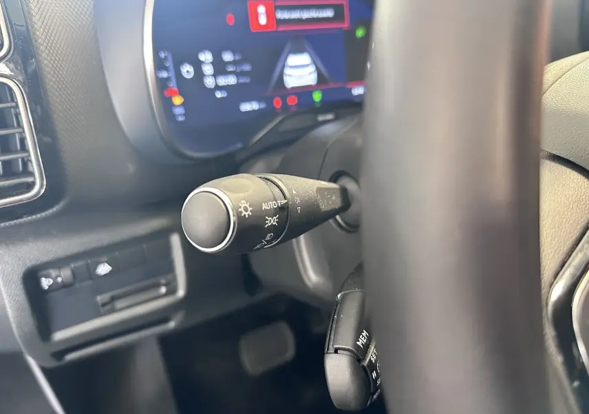Gros plan sur la commande des phares et le volant cuir du Citroën C5 Aircross 2024, tableau de bord numérique en arrière-plan.