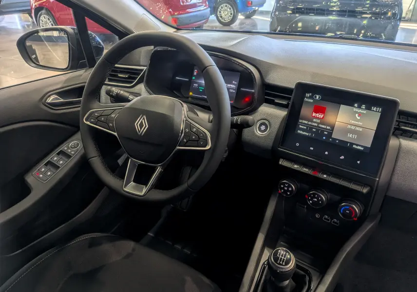 Vue intérieure de face du poste de conduite de la Renault Clio V avec volant, écran tactile et levier de vitesses manuel.