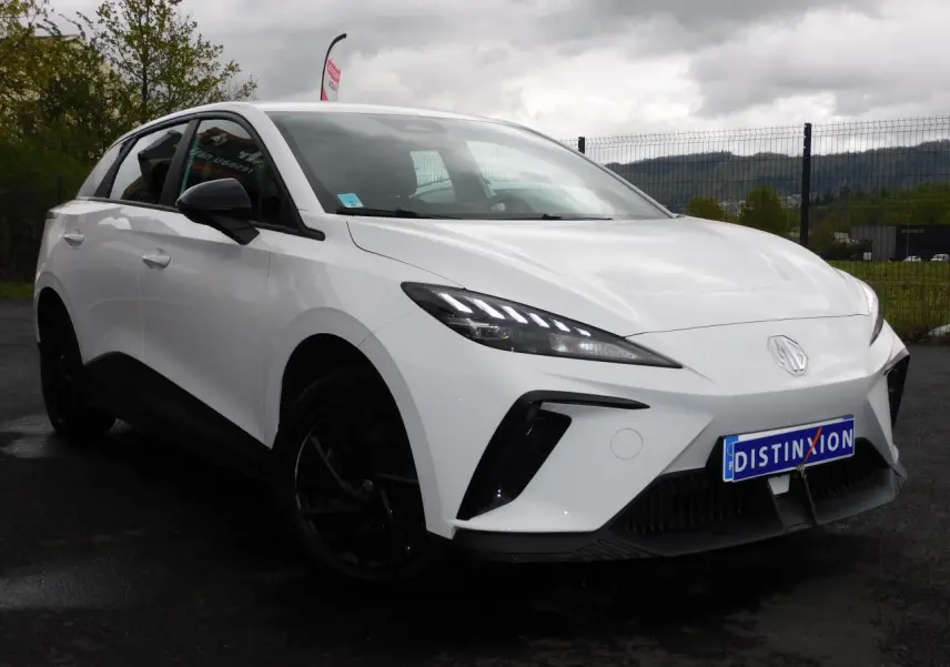 Vue 3/4 avant d'une MG4 électrique blanche avec jantes noires et feux LED distinctifs sur un parking extérieur.