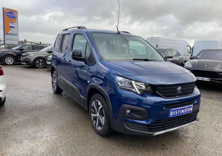Peugeot Rifter GT bleu métallisé en 3/4 avant droit sous ciel nuageux sur parking humide.