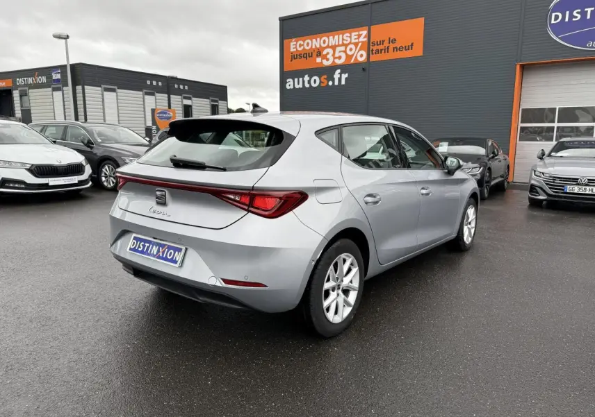 Vue 3/4 arrière droite d'une SEAT Leon 2022 en argent urban, stationnée devant un concessionnaire sous un ciel nuageux.