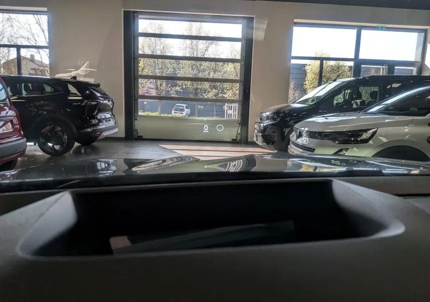 Vue intérieure depuis le tableau de bord montrant le Renault Austral gris schiste avec toit noir en 3/4 avant droit dans un garage lumineux.