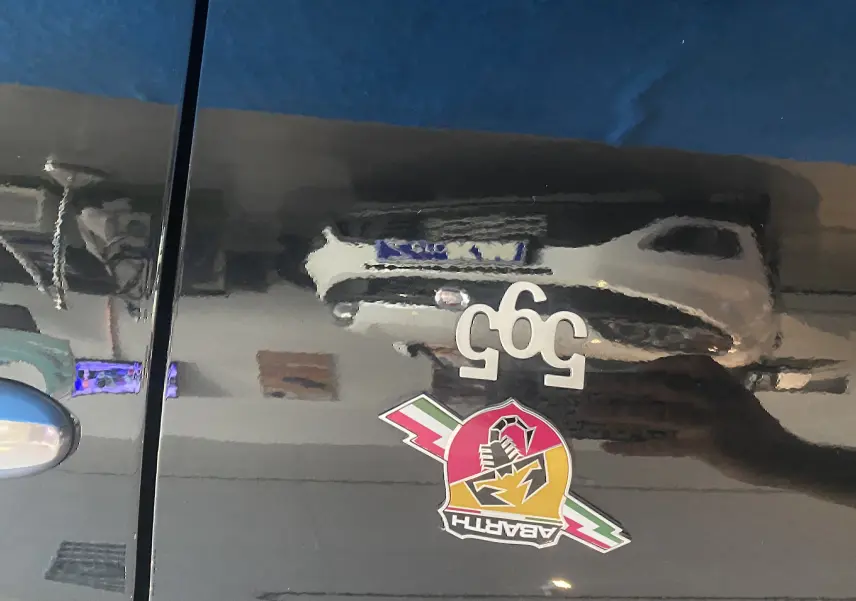 Gros plan sur la porte noire d'une Abarth 595 avec logo scorpion et badge 595 blanc visible en reflet.