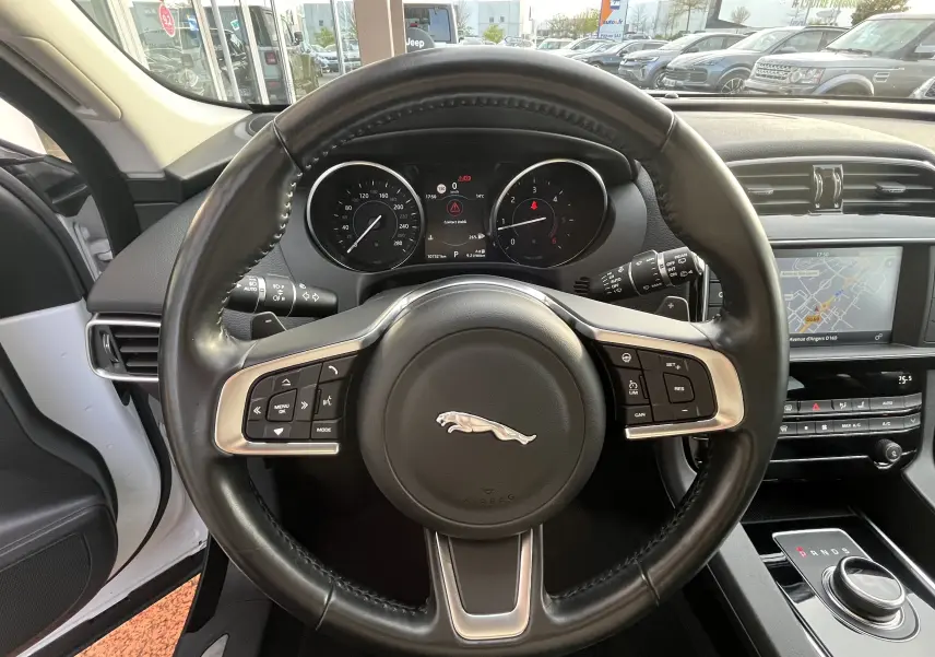Vue intérieure centrée sur le volant cuir noir du Jaguar F-PACE blanc 2018 avec tableau de bord et écran GPS visible.
