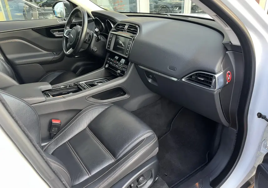 Vue intérieure côté conducteur du Jaguar F-PACE blanc 2018, avec sellerie cuir noir et tableau de bord moderne.
