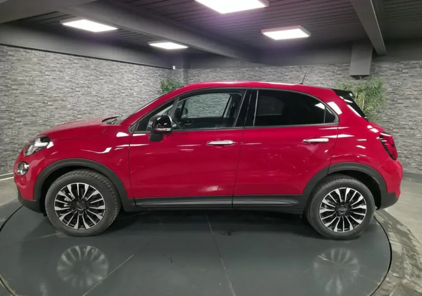 Profil droit d'un Fiat 500X rouge Passione 2024 avec jantes alliage 17'' et vitres surteintées en intérieur showroom.