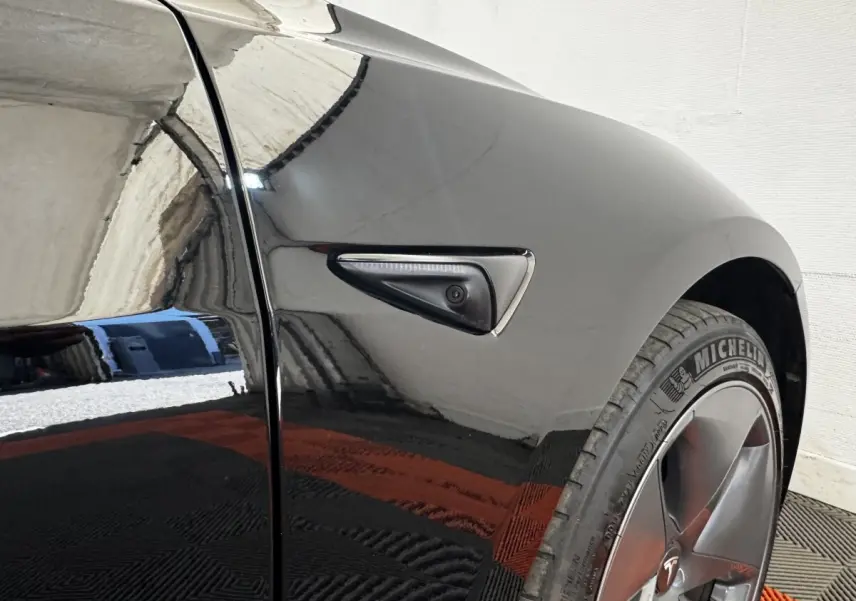 Gros plan sur l'aile avant droite noire d'une Tesla Model 3 2023 avec caméra latérale intégrée et jante grise.