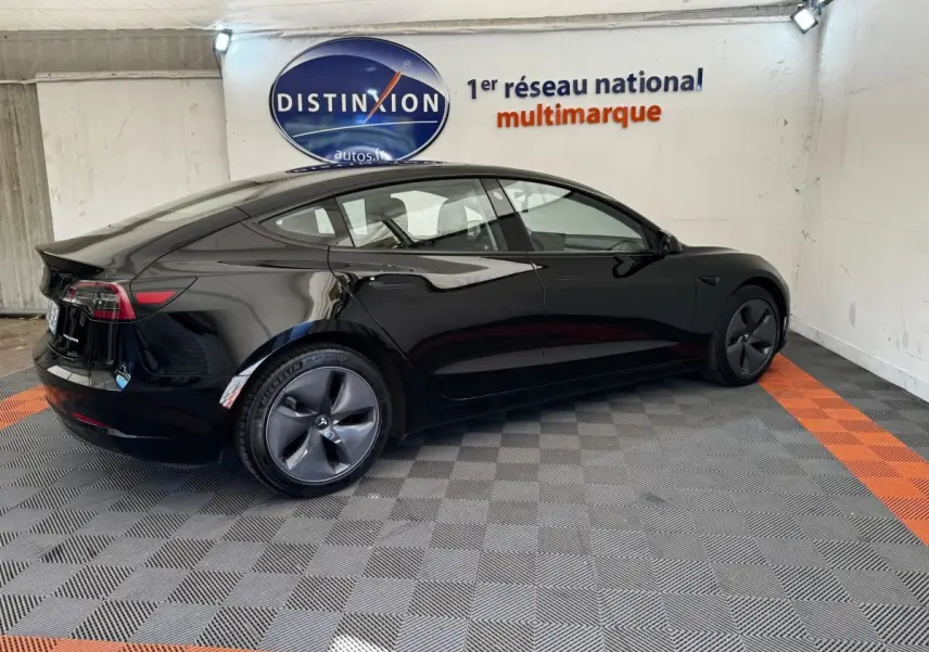 Tesla Model 3 Grande Autonomie noir, vue 3/4 arrière côté gauche dans un showroom avec sol gris et orange.