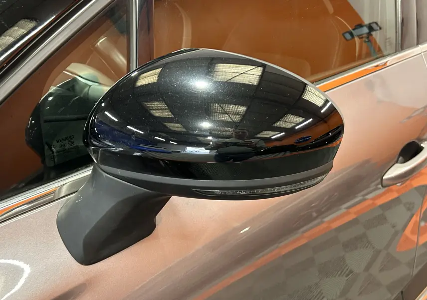 Rétroviseur latéral noir brillant sur côté gauche d’un Renault Captur gris avec intérieur marron visible.