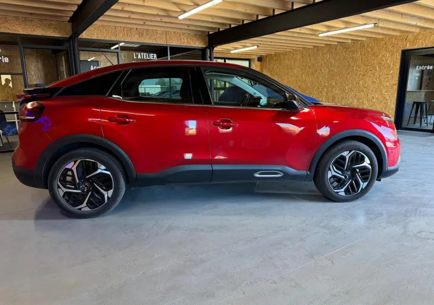 Vue de profil côté gauche d'une Citroën C4 X rouge avec jantes noires et détails noirs sous un abri en bois.