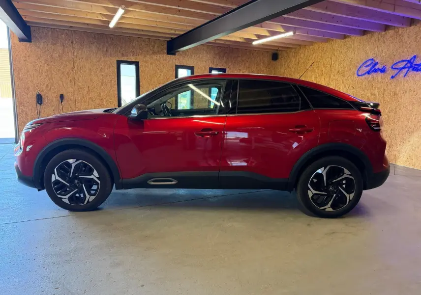 Vue de profil côté gauche d'une Citroën C4 X rouge brillant avec jantes noires et toit noir dans un garage.