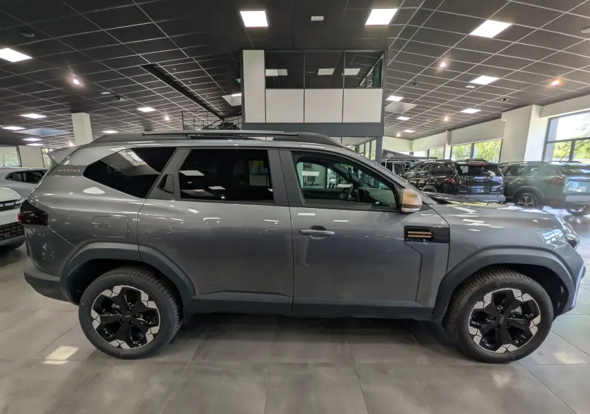 Vue de profil côté gauche du Dacia Bigster Hybrid-G 150 4x4 gris schiste avec jantes biton noires en showroom.