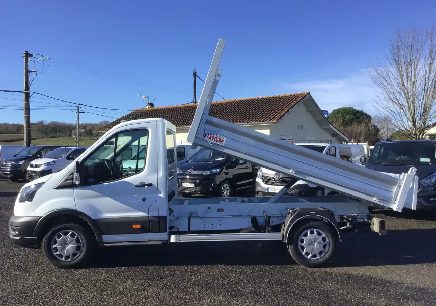 Vue de profil côté gauche d'un Ford Transit blanc avec benne basculante levée sur un parking extérieur.