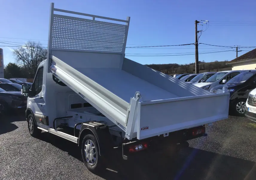 Vue 3/4 arrière droite d'un Ford Transit blanc avec benne relevée et grille de protection arrière sur parking.