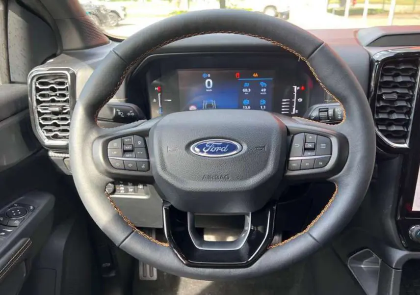 Vue rapprochée du volant noir avec surpiqûres orange du Ford Ranger 3.0 V6 240 Wildtrak Plus 2025, tableau de bord numérique visible.