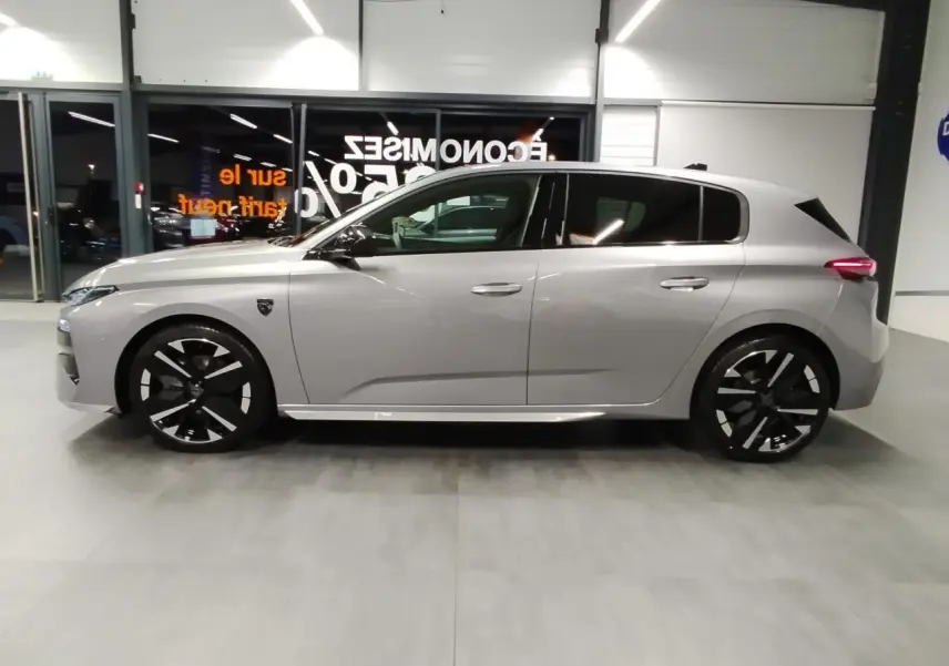 Peugeot 308 Hybrid gris Artense vue de profil côté gauche, jantes alliage noires et toit noir brillant visible en intérieur showroom.