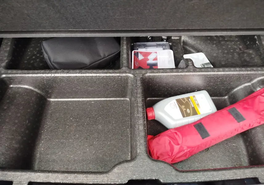 Compartiment de rangement sous le coffre du Kia Sportage bleu, avec trousse de secours rouge et bidon d'huile visible.