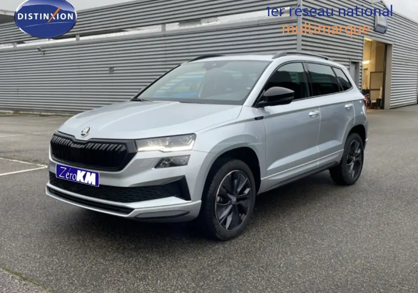 Vue 3/4 avant d'un Skoda Karoq 2025 gris argent métal avec calandre noire et jantes foncées, stationné devant un bâtiment industriel.