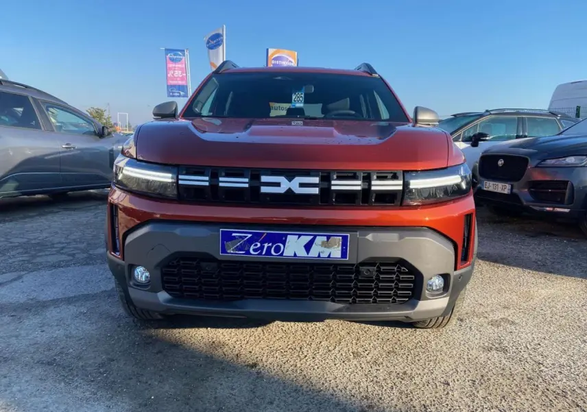 Vue de face d'un Dacia Duster 2024 brun terracotta avec calandre marquée et plaques "Zéro KM" sur un parking extérieur.
