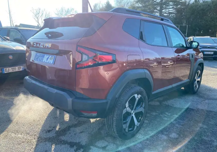 Vue 3/4 arrière droite du Dacia Duster 2024 brun terracotta avec jantes alliage et barres de toit noires.