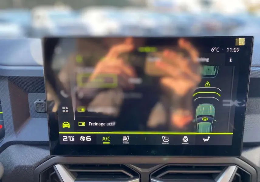 Écran tactile central du Dacia Duster 2024 affichant l'aide au freinage et la climatisation, vue intérieure frontale.