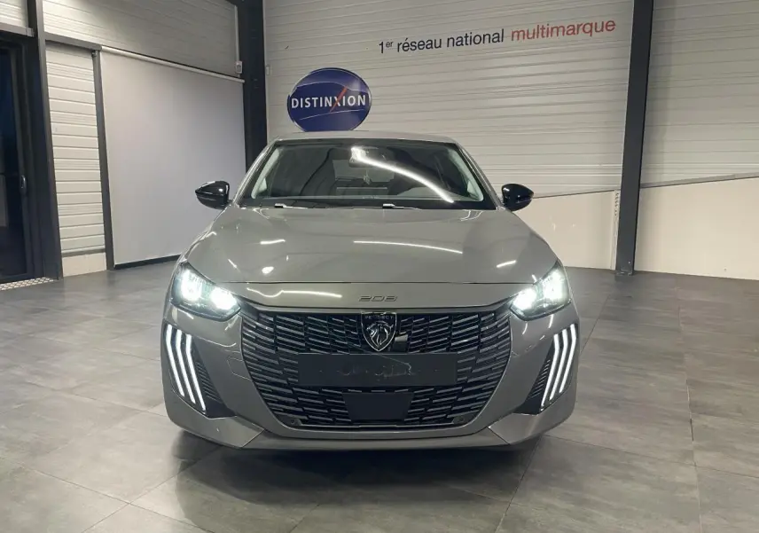 Vue avant d'une Peugeot 208 gris selenium avec phares LED allumés et calandre noire brillante en intérieur showroom.