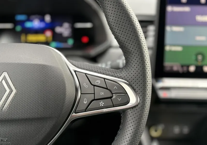 Gros plan sur le volant perforé noir du Renault Captur 2025 avec commandes intégrées et écran numérique flou en arrière-plan.