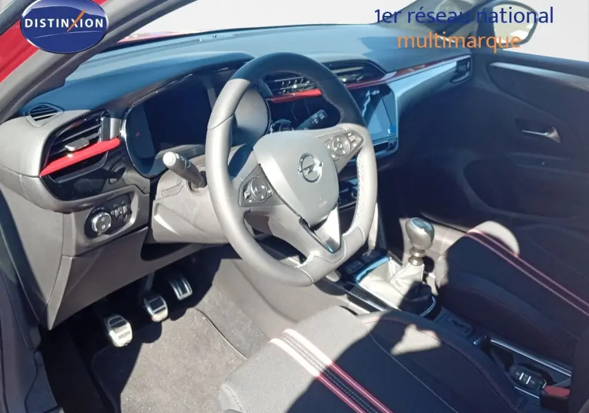 Vue intérieure de l'Opel Corsa 1.2 Turbo 2023 montrant le volant, la boîte manuelle et les sièges noirs avec surpiqûres rouges.