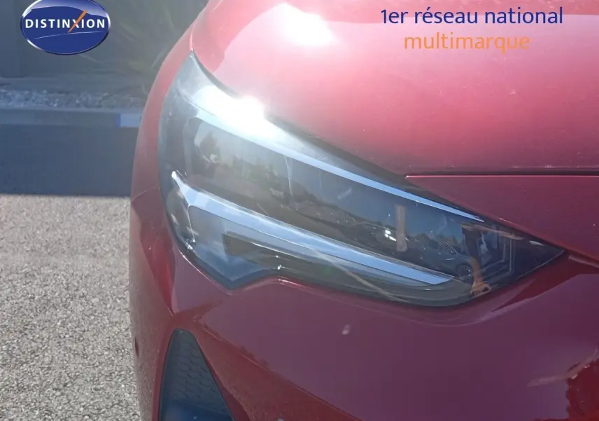 Gros plan sur le phare avant gauche d'une Opel Corsa 2023 rouge métal avec toit noir, sous un soleil éclatant.