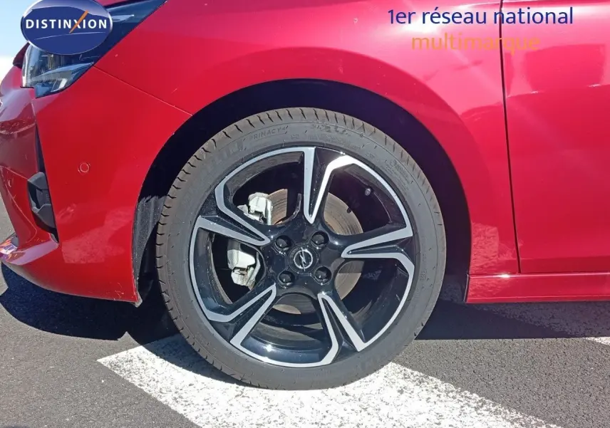 Gros plan sur la roue avant gauche d'une Opel Corsa rouge métal avec jante noire et détails argentés.