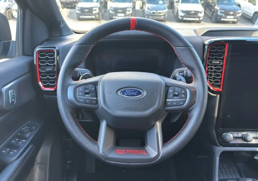 Vue intérieure centrée sur le volant noir du Ford Ranger Raptor 2024 avec surpiqûres rouges et aérateurs rouges.