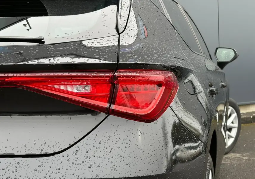 Vue arrière droite d'une SEAT Leon 2024 noir minuit, avec gouttes de pluie sur la carrosserie et feux arrière allumés.