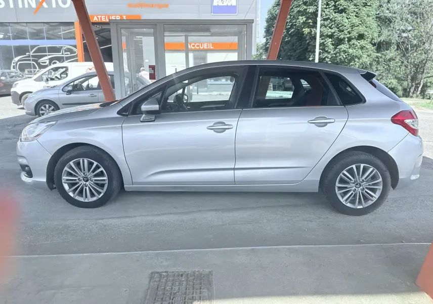 Vue de profil côté gauche d'une Citroën C4 gris métallisé, 5 portes, garée sous un abri avec fond urbain.