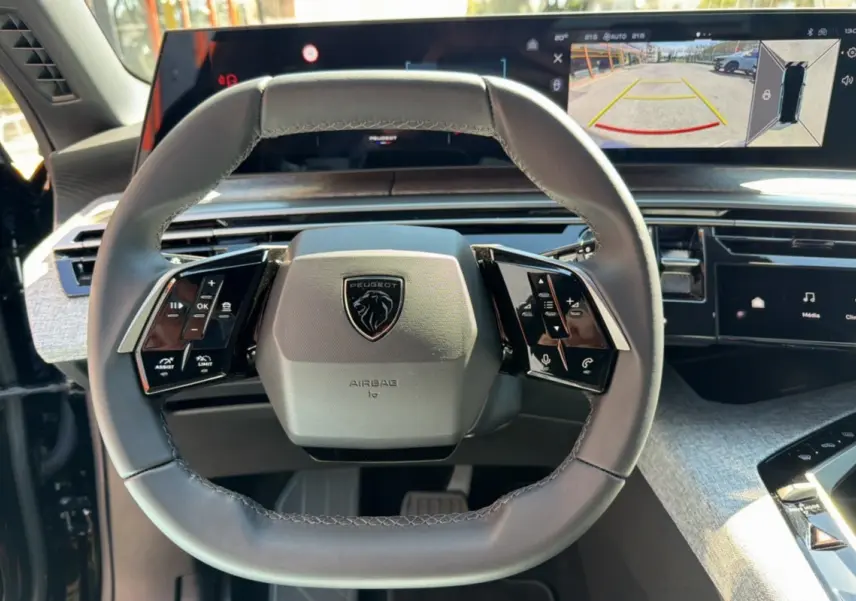 Vue rapprochée du volant cuir multifonctions gris du Peugeot 5008 Hybrid 145 avec écran panoramique affichant la caméra 360° en arrière-plan.