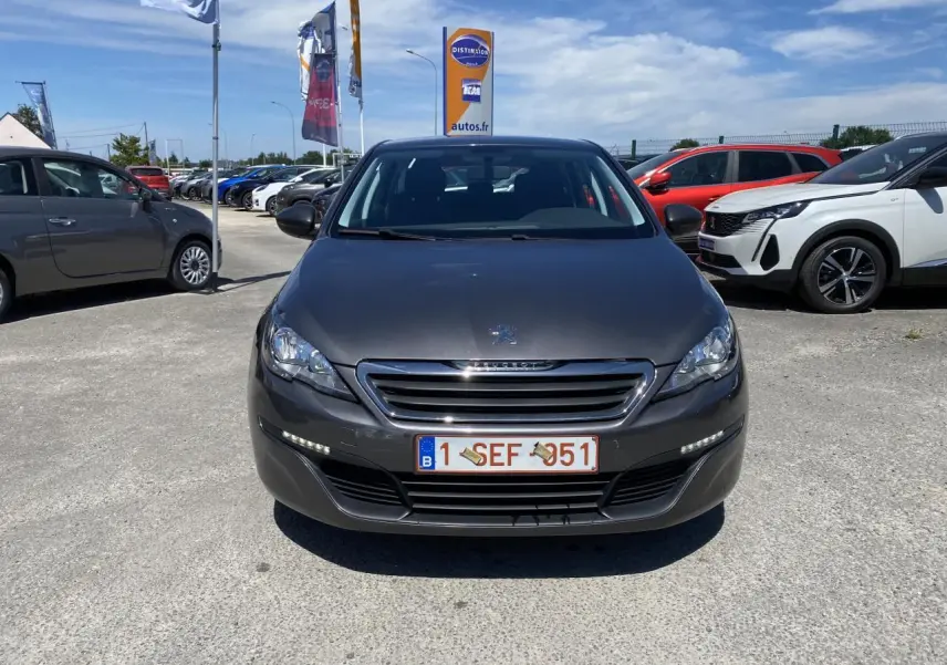 Vue frontale d'une Peugeot 308 SW gris clair garée en extérieur sous un ciel bleu avec d'autres véhicules autour.
