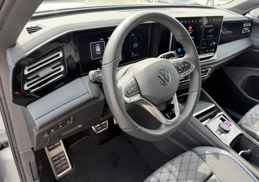 Vue intérieure côté conducteur du Volkswagen Tiguan 2025, volant cuir noir, tableau de bord digital et sièges gris à surpiqûres bleues.