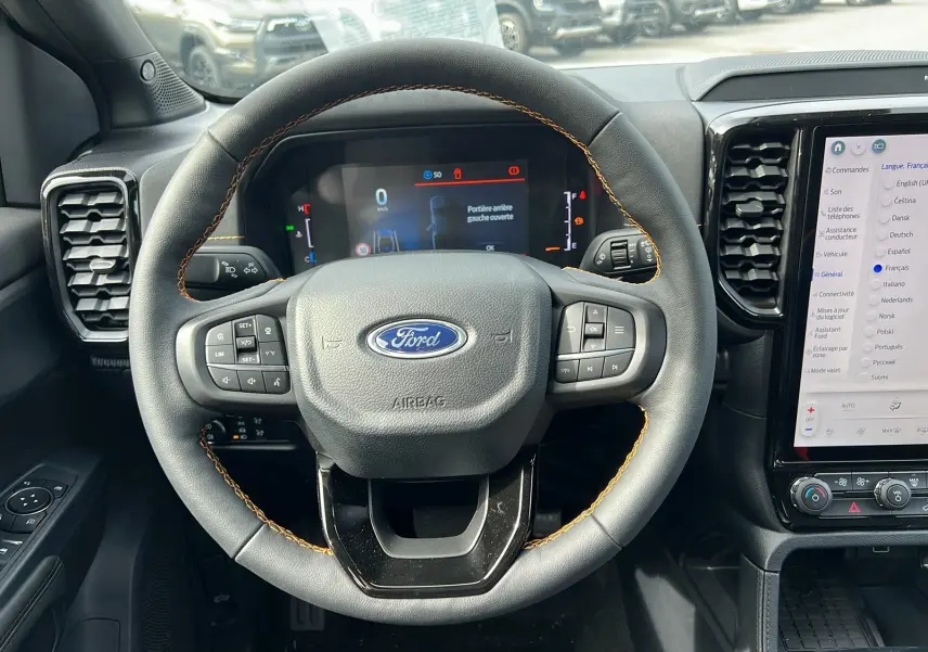 Vue rapprochée du volant cuir noir avec surpiqûres orange du Ford Ranger 2025, tableau de bord digital et écran tactile visible.