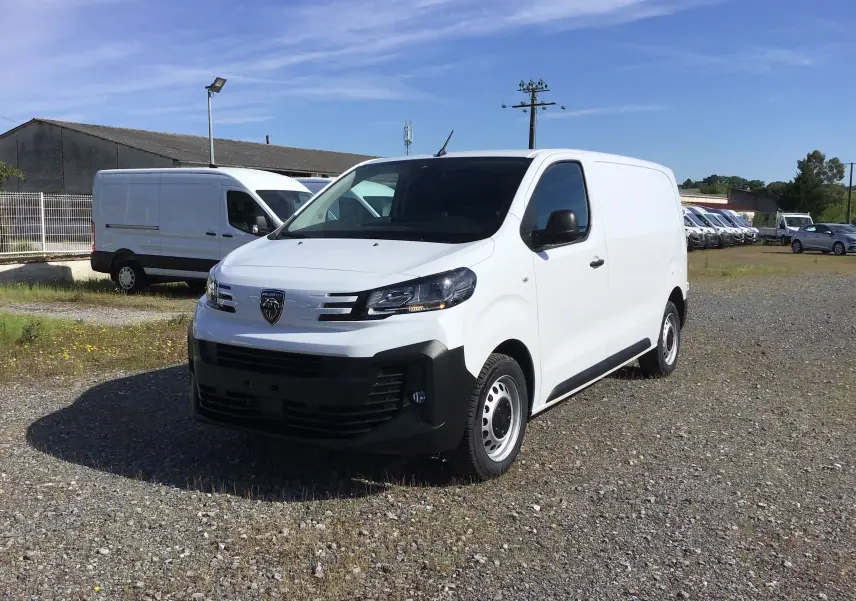 Fourgon blanc Peugeot Expert vu en 3/4 avant droit sur un terrain gravillonné avec plusieurs véhicules en arrière-plan.
