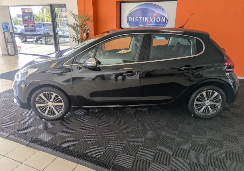 Profil côté gauche d'une Peugeot 208 Allure noire de 2019, cinq portes, avec jantes alliage bicolores, en intérieur showroom.